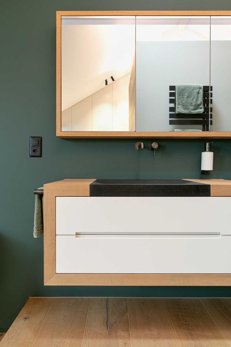 Dieser Unterbau mit umlaufenden Elementen in Eiche harmoniert elegant mit dem eingelassenen Steinbecken. Die spritzlackierten Fronten mit eingefrästen Griffprofilen sorgen für eine klare, ruhige Optik. Der Badschrank mit umlaufendem Eichen-Rahmen, Doppelspiegeltüren sowie integrierter indirekter Beleuchtung runden das Ensemble funktional und hochwertig ab. Oesch Innenausbau AG, www.oesch.ch