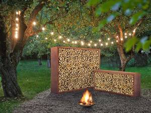 Eine Feuerschale inmitten eines Waldstücks, dazu ein Holzlager, beleuchtet und in Szene gesetzt. Sager steht für hochwertige Gartenobjekte, die individuell auf Mass angefertigt werden oder auf Standardgrössen basieren. Sager Gartengalerie AG.