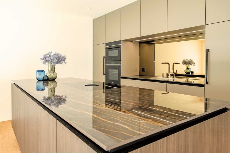 Tiefe und Helligkeit: Diese stilvolle Küche vereint modernes Design mit funktionalen Elementen. Die Fronten in Achatgrau der «X-Line»-Griffleiste wirken elegant und zeitlos. Ein Highlight ist die polierte Keramik­abdeckung «Noir Desir», die mit ihrem dezenten Glanz edle Akzente setzt. Eindrucksvoll ist die bronzefarbene Spiegelrückwand, die Licht reflektiert und dem Raum Helligkeit und Tiefe verleiht. 

Hans Eisenring AG, www.eisenring-kuechenbau.ch